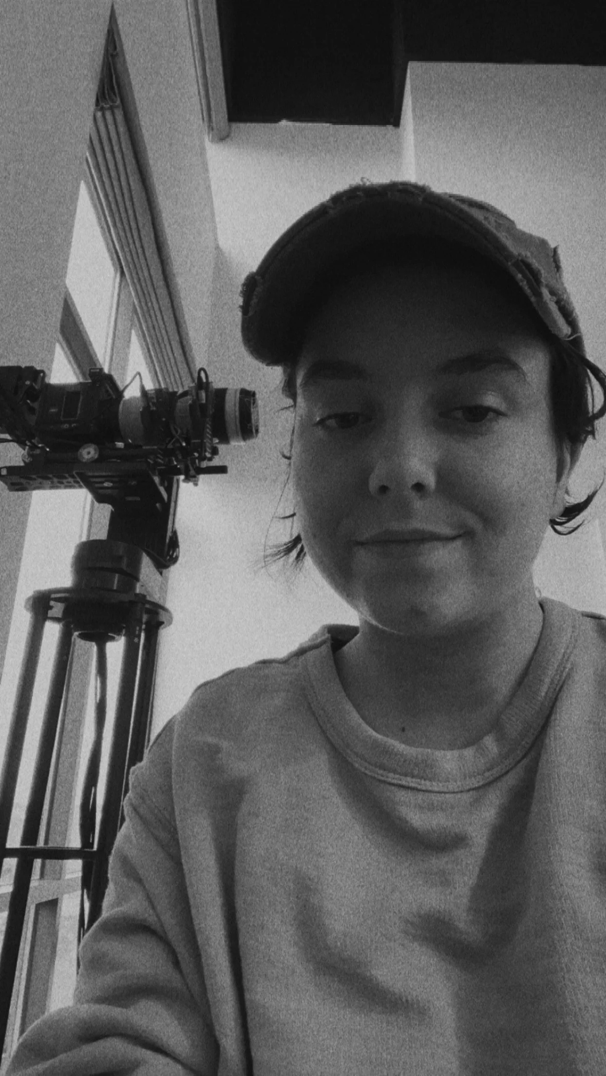 A young person with short hair wearing a cap, taking a selfie indoors next to a camera mounted on a tripod.