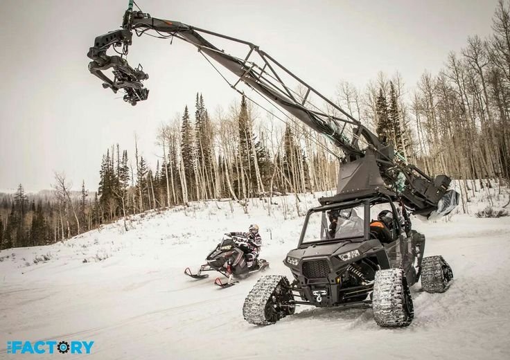 A snow-covered landscape with a person riding a snowmobile and a large camera crane vehicle.