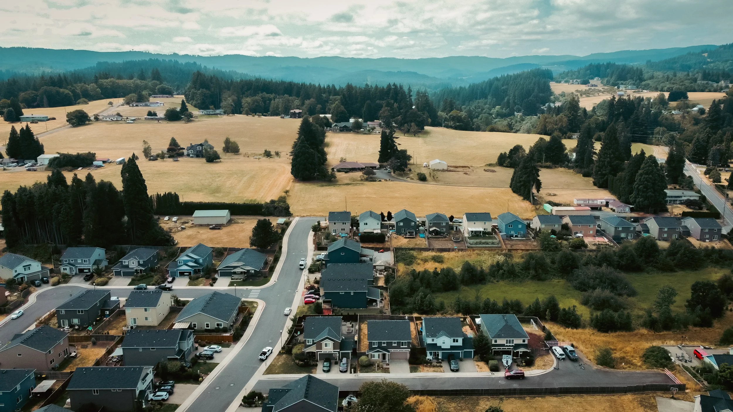 Aerial view of a suburban neighborhood with houses, trees, and open fields, with hills and mountains in the background under a partly cloudy sky.