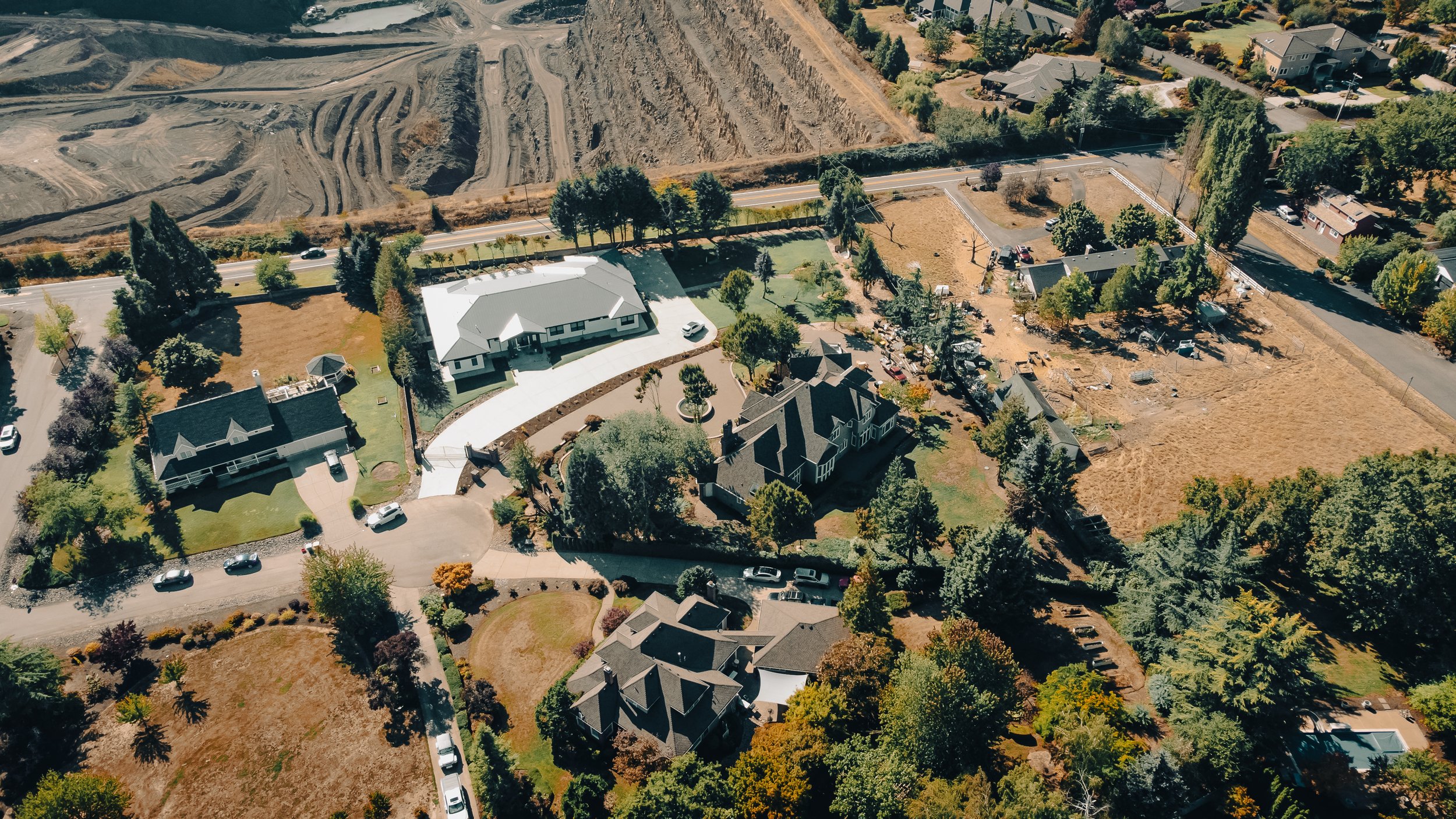 Aerial view of a residential neighborhood with large houses, trees, and a construction site or cleared land at the top of the image.