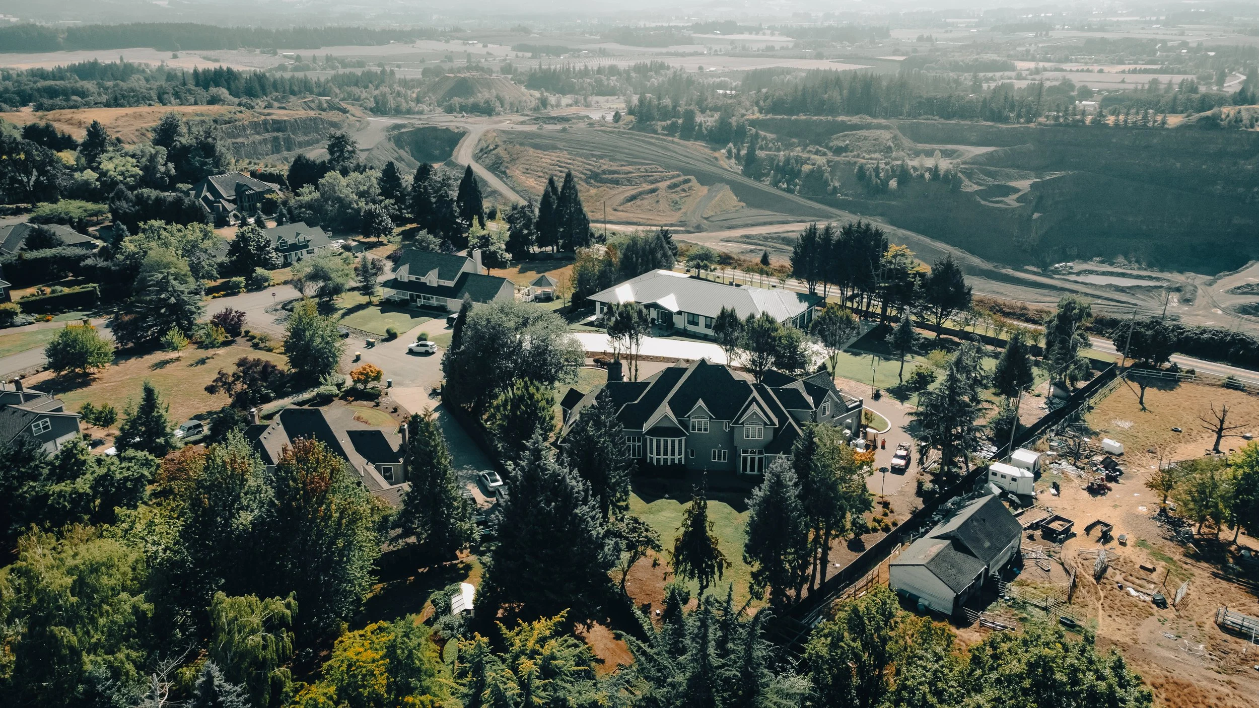 Aerial view of a residential neighborhood with houses, trees, and yards, with a backdrop of rolling hills and a quarry in the distance.