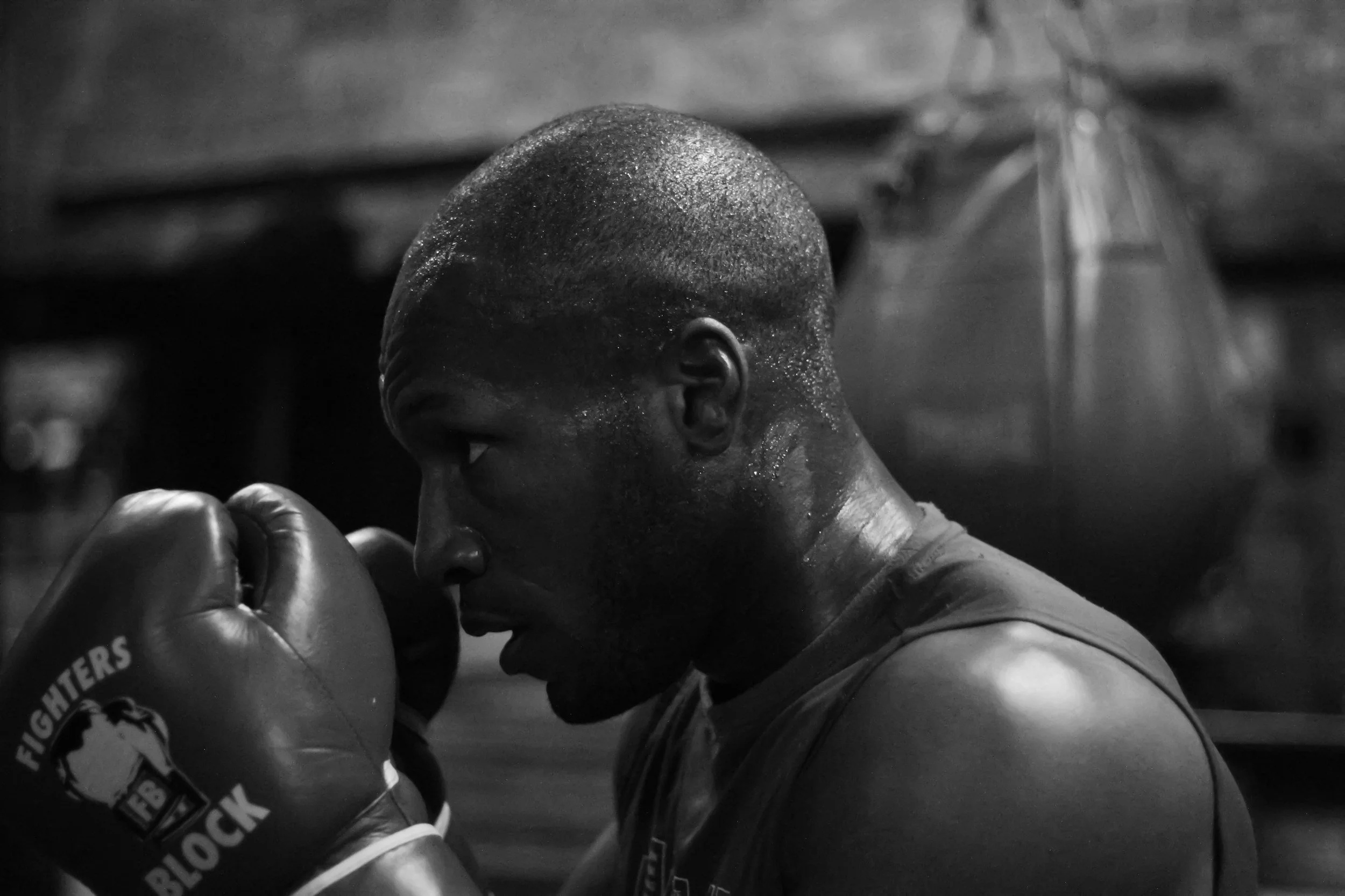 A male boxer in a training gym, wearing boxing gloves, appears to be focusing or preparing for a workout.