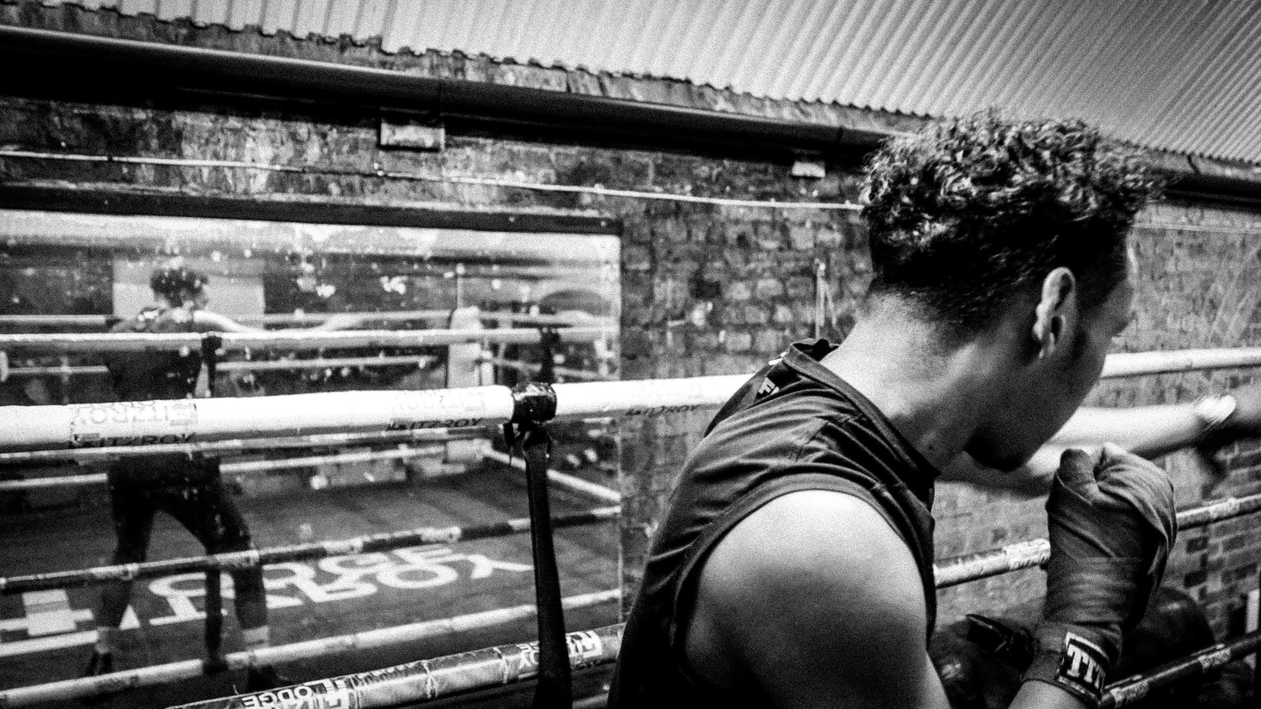 A person with curly hair and muscular arms in a boxing gym, wearing boxing gloves and preparing to punch a focus mitt held by a trainer. The gym has brick walls and a mirror reflecting other individuals training.
