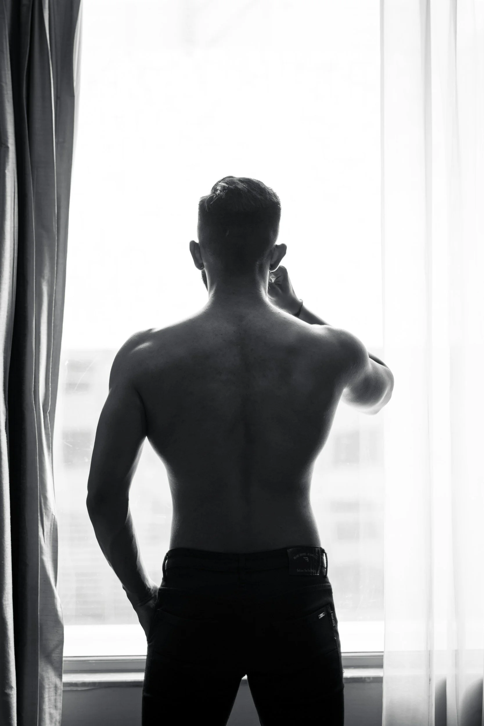 A shirtless man with tattoos on his back and arms stands in front of a large window with sheer curtains, looking out.