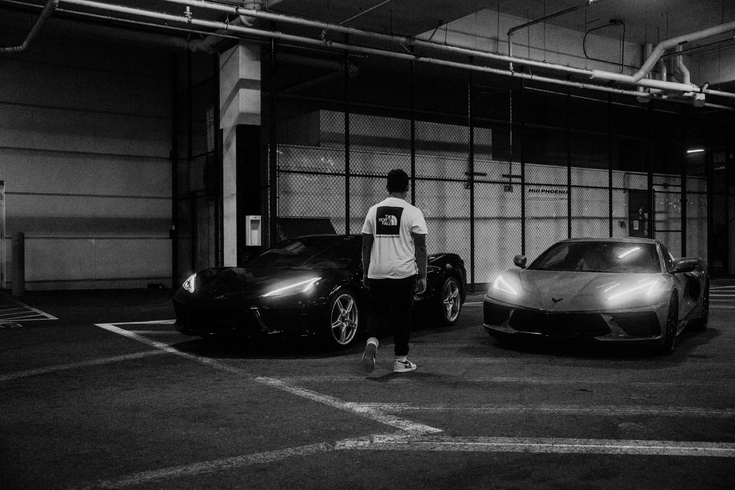 A person with short hair wearing sneakers and a white T-shirt with The North Face logo walking through a parking lot at night. The lot contains black and gray sports cars, and a chain-link fence is visible in the background with large industrial pipe