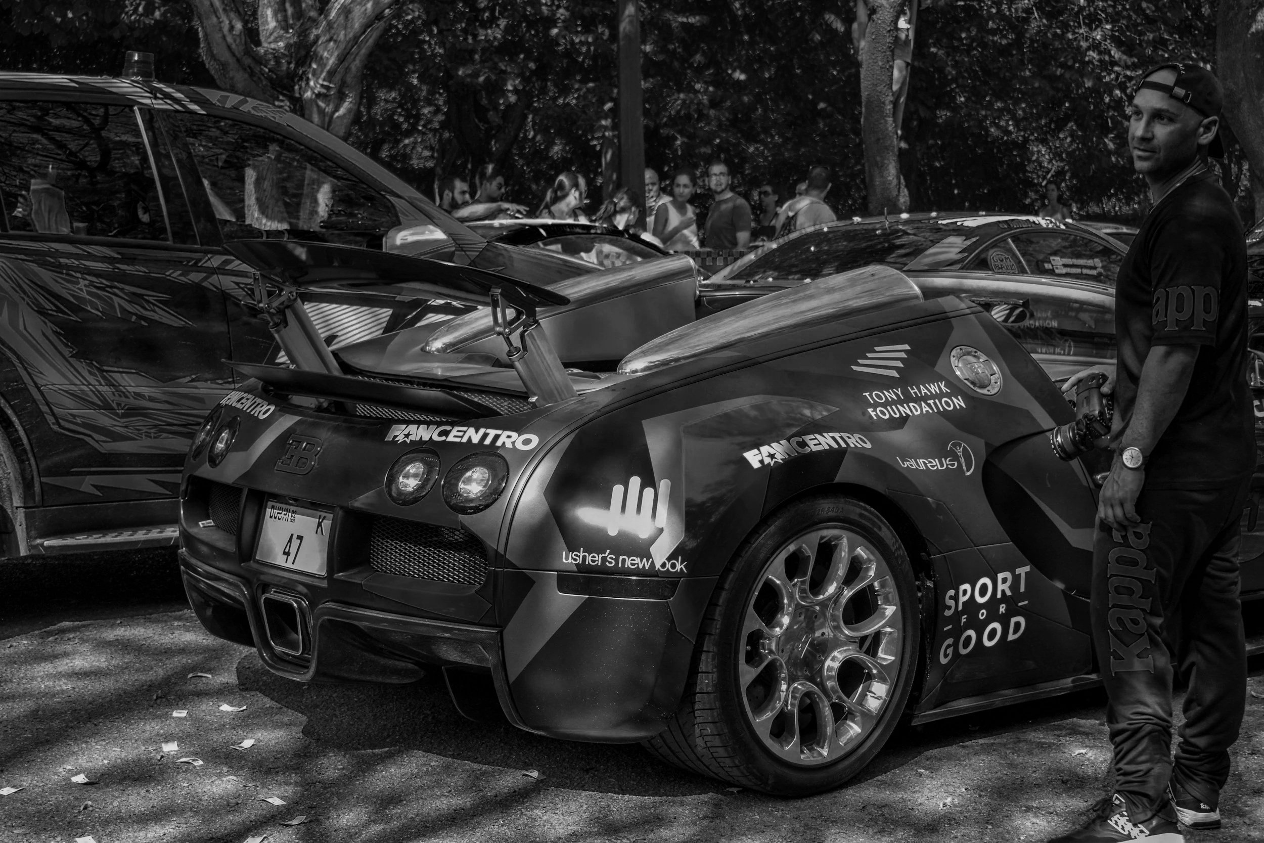 Black and white photo of a race car with sponsorship logos, a person holding a camera, and people in the background at an outdoor event.