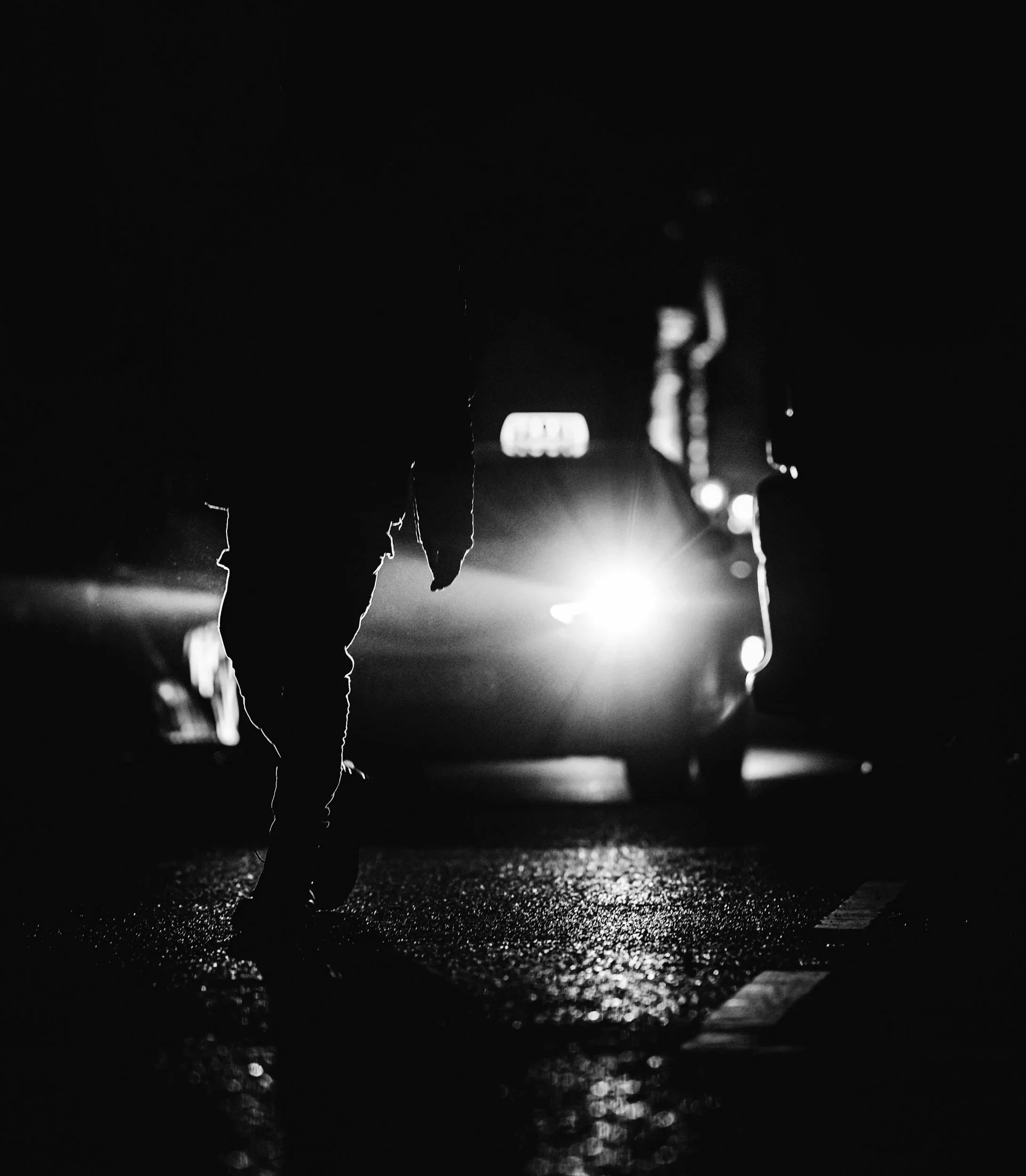 Silhouette of a person walking in the rain at night, with bright light behind them creating a glow and reflective surfaces showing rain drops.
