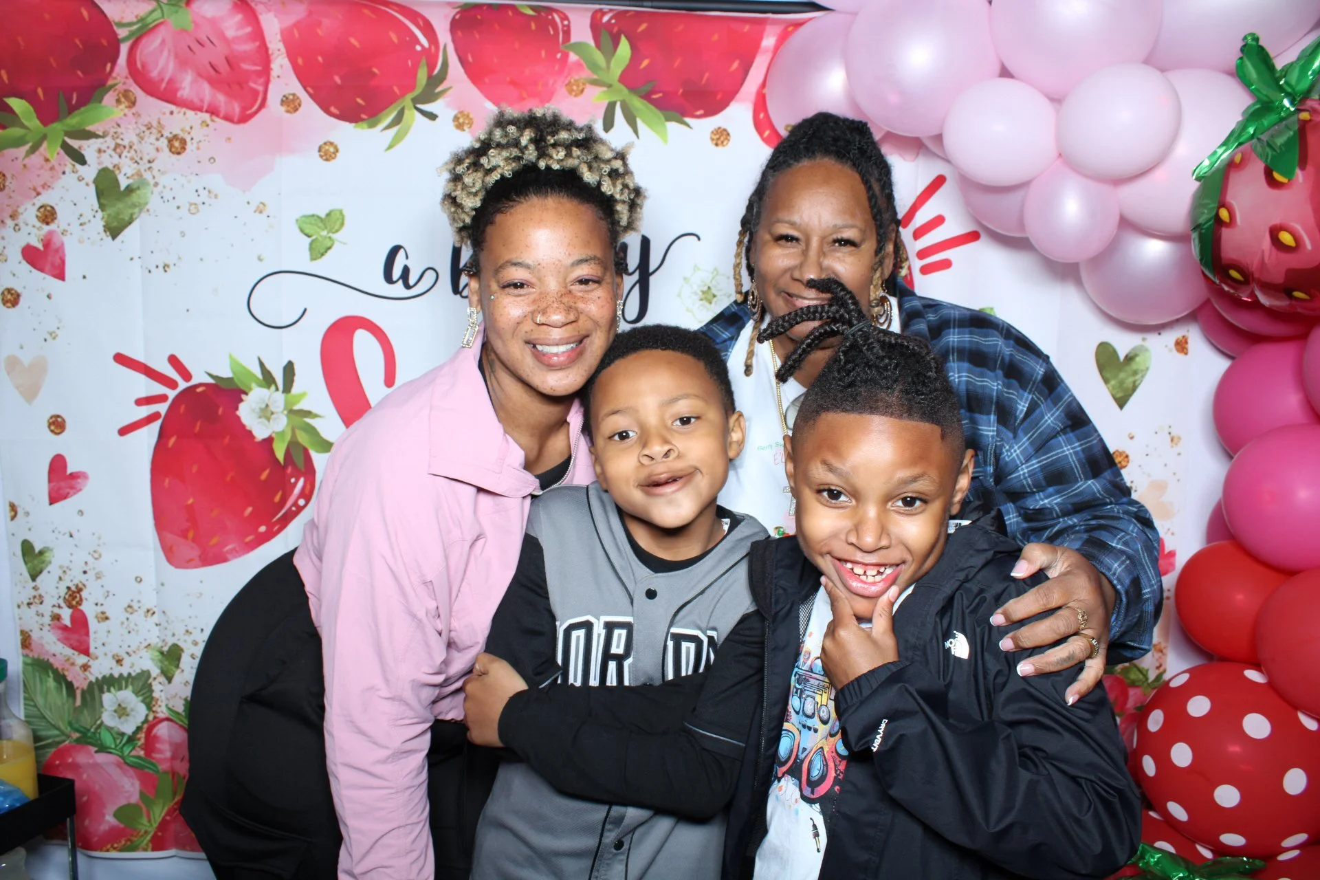 Photobooth at a baby shower. A smiling woman with light brown hair and a pink shirt, a woman with dark curly hair and a blue plaid shirt, and two young boys, one with short hair and a gray jacket, and the other with short hair and a black jacket.