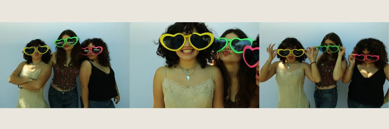 Four women wearing colorful heart-shaped sunglasses posing for a photo against a plain background.