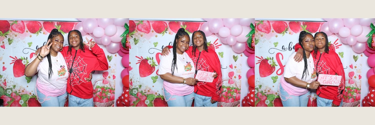 Three women at a strawberry-themed celebration or event with pink balloons, holding signs that say 'It's a Girl' and 'Good Morning'.