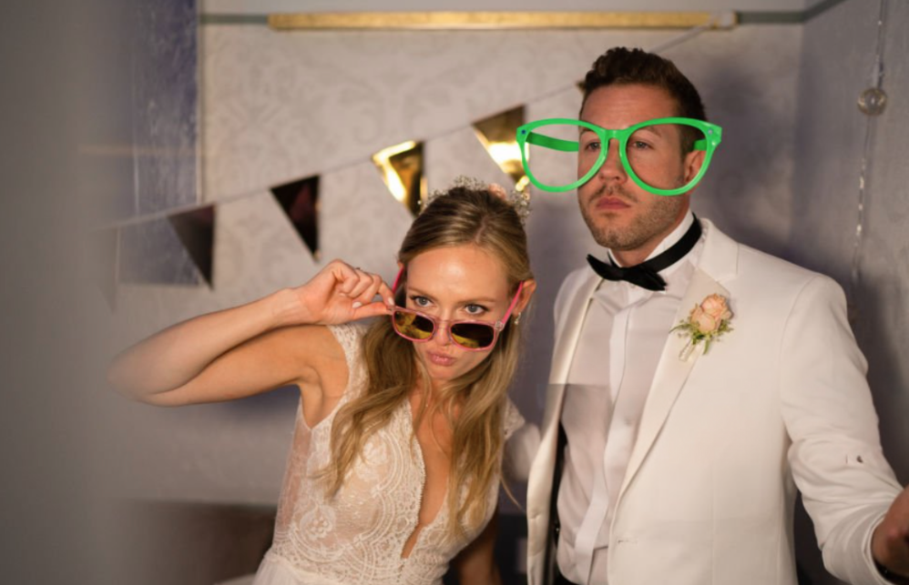 Photobooth at a wedding. A woman and a man in formal attire wearing oversized novelty glasses at a party or celebration. The woman is holding sunglasses and making a playful face, while the man is wearing large green glasses and looking serious.