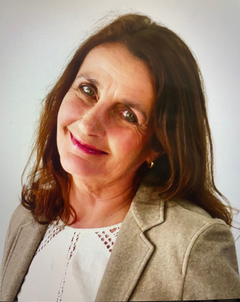 A woman with shoulder-length brown hair and light skin, smiling and wearing a beige blazer over a white top, standing against a plain light-colored background.