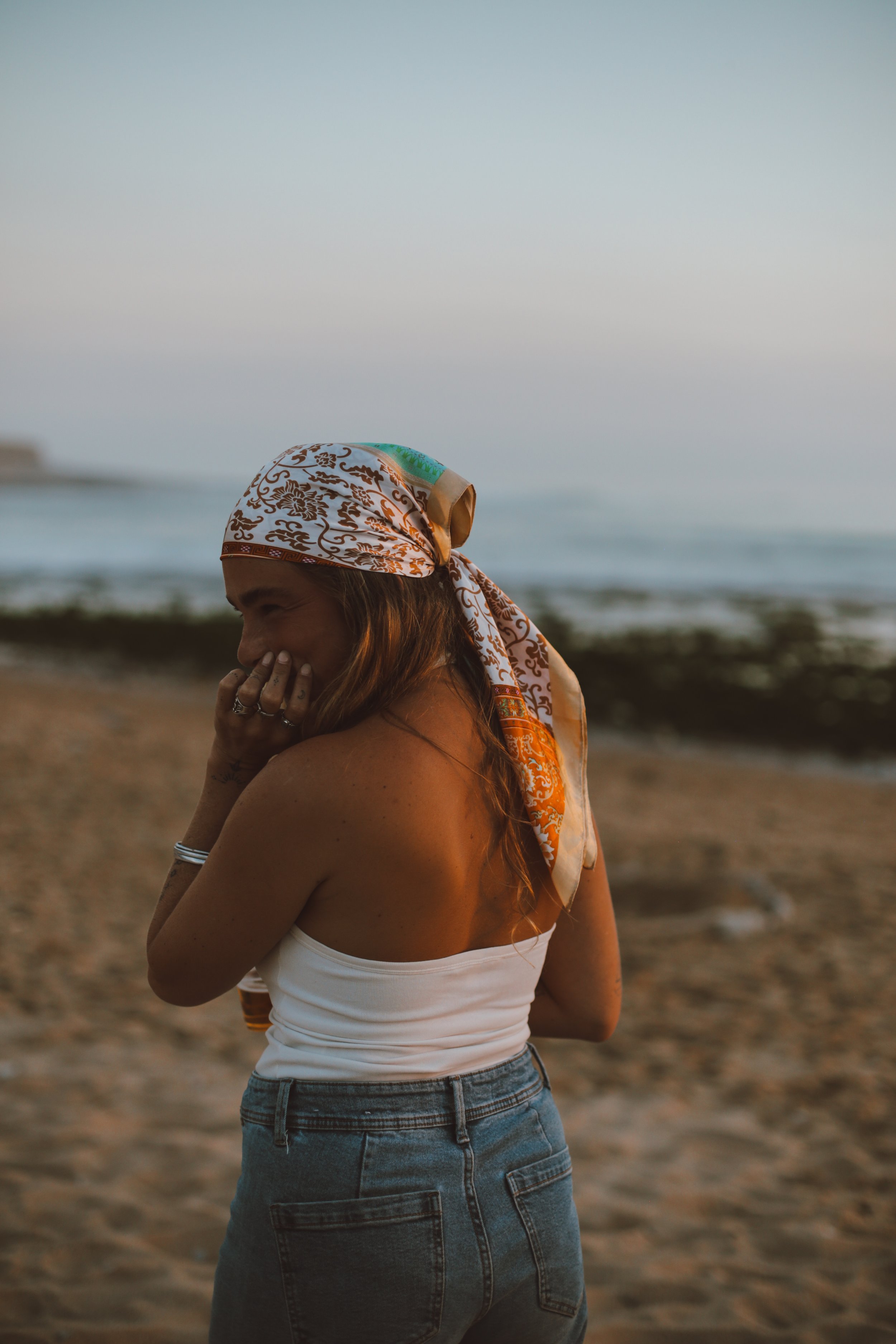 A woman with long hair wearing a white tank top, denim shorts, and a patterned headscarf, standing on the beach at sunset, smiling and touching her face.