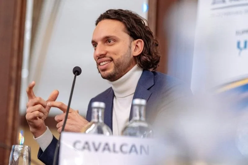 Man speaking at a panel, gesturing with his hand, sitting behind a table with water bottles, with a sign that says "CAVALCAN" in front of him.