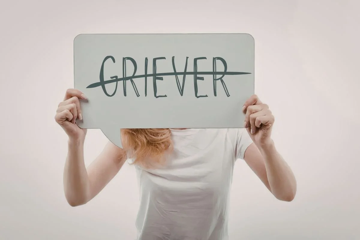 Woman hiding behind a signed with the label "griever" with the word crossed out.