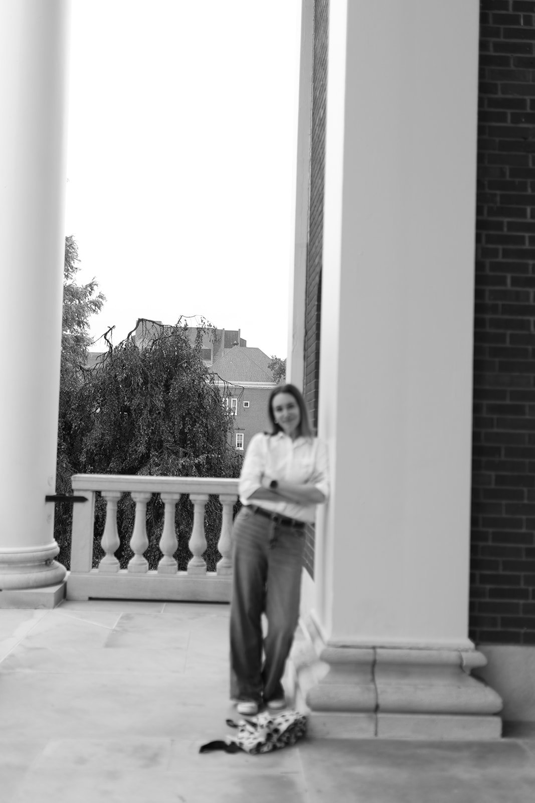 A woman leaning against a column on a balcony, with a railing, trees, and buildings in the background, captured in black and white.