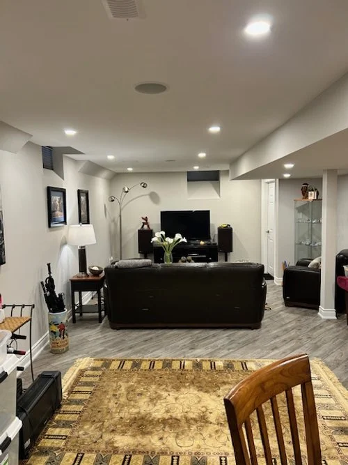 Living room with a black leather sofa facing a television, a vase of white flowers on a table, a brown area rug, and various furniture pieces and decorations.