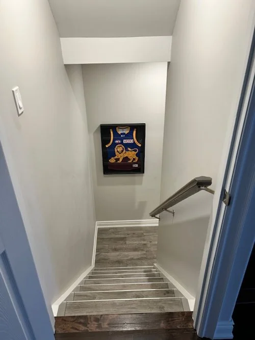 A staircase with gray wood steps and white risers, descending into a small landing with white walls and a railing on the right side. There's a framed picture of a lion and shoes hanging on the wall at the bottom of the stairs.