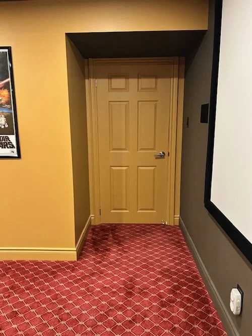 A beige door with a silver handle located at the end of a hallway, with a red patterned carpet on the floor and yellow and gray walls.