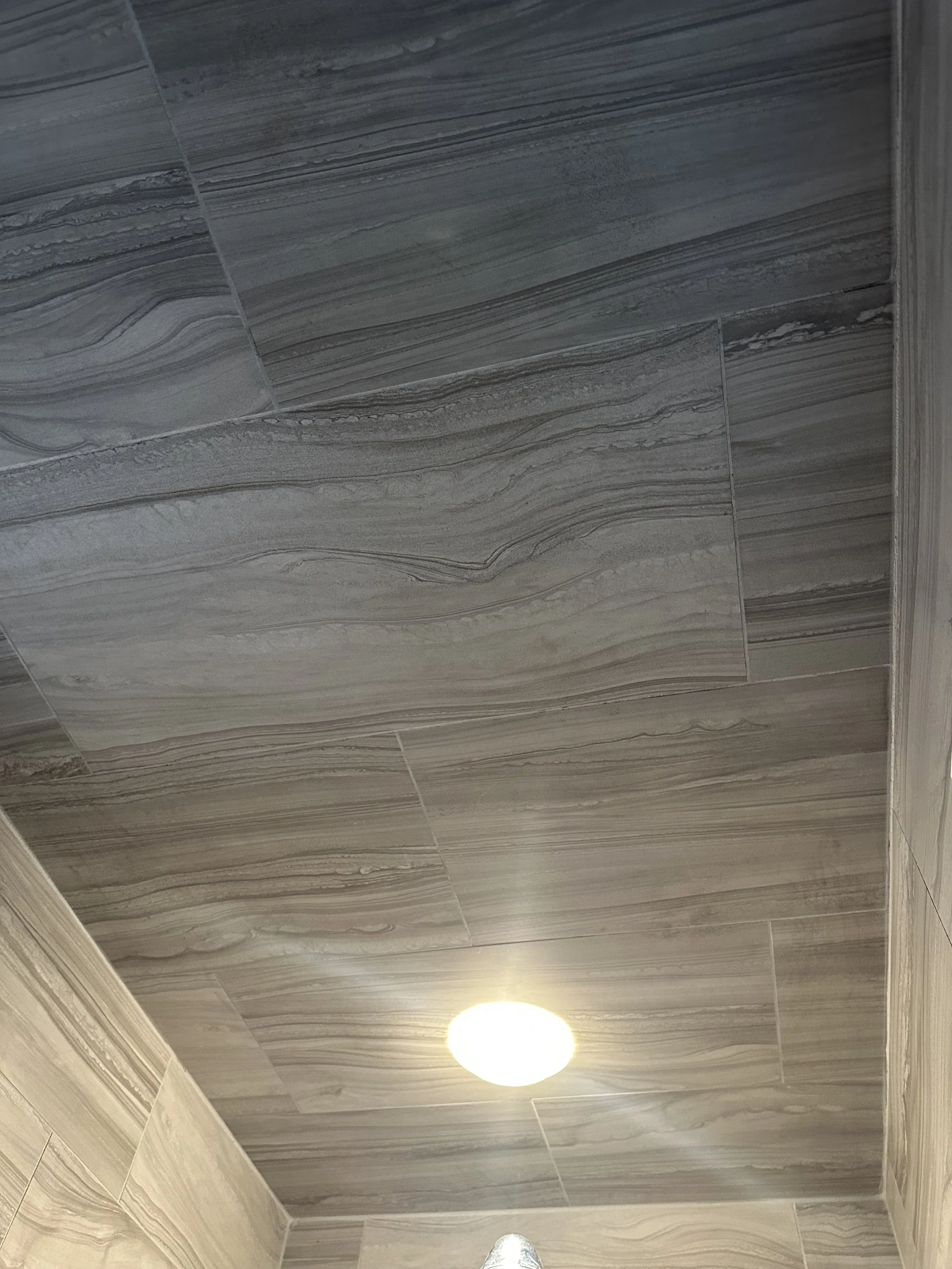 Ceiling of a bathroom with large grey marble tiles and a round recessed light fixture.