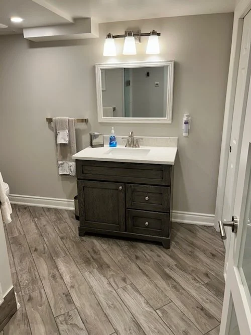 Bathroom with a dark vanity, white countertop, rectangular mirror, three-light fixture, gray wood-look flooring, towel rack with towels, soap dispenser, and a partially visible shower door.