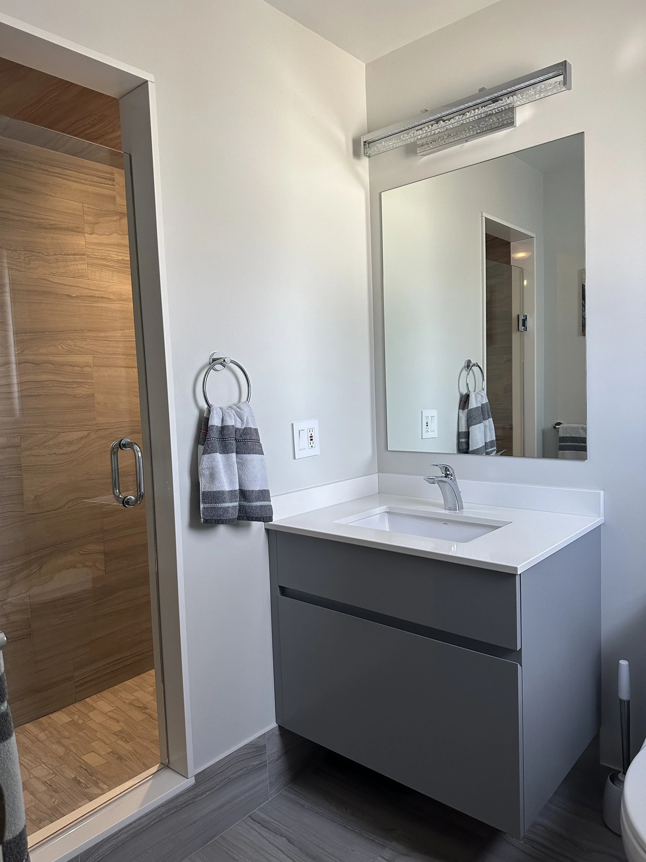 Modern bathroom with a white vanity, grey cabinet, large mirror, and a glass-enclosed shower with wood-look tiles.