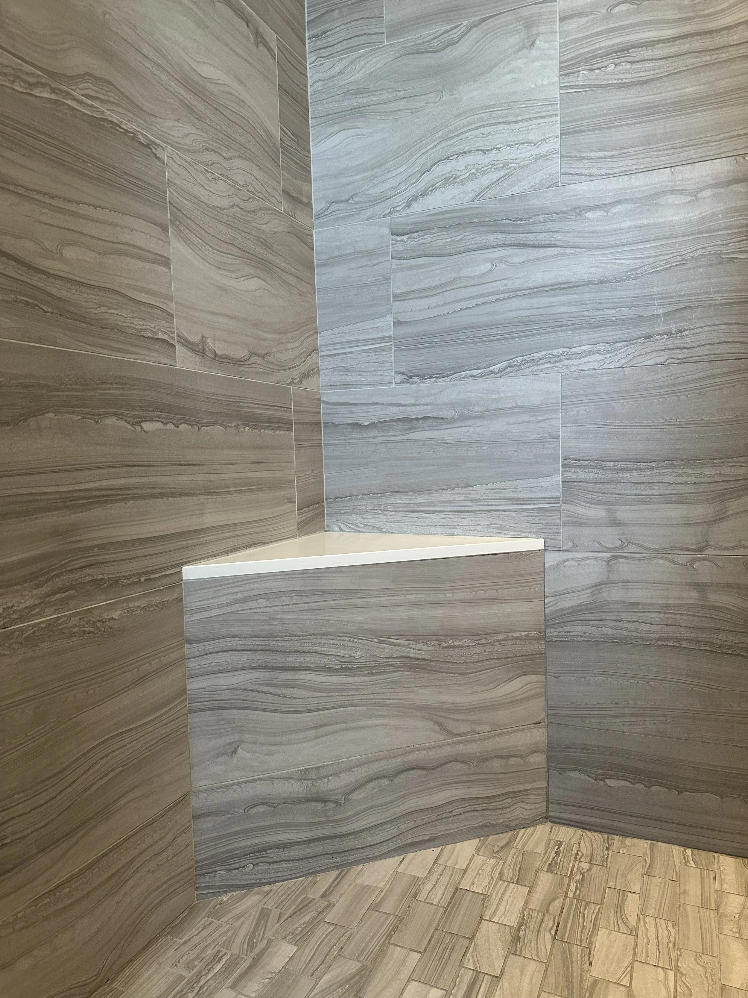Close-up of a corner in a bathroom with marble-patterned tiles on the walls and a built-in ledge or shelf with a white top. The flooring consists of small rectangular tiles in a beige tone.