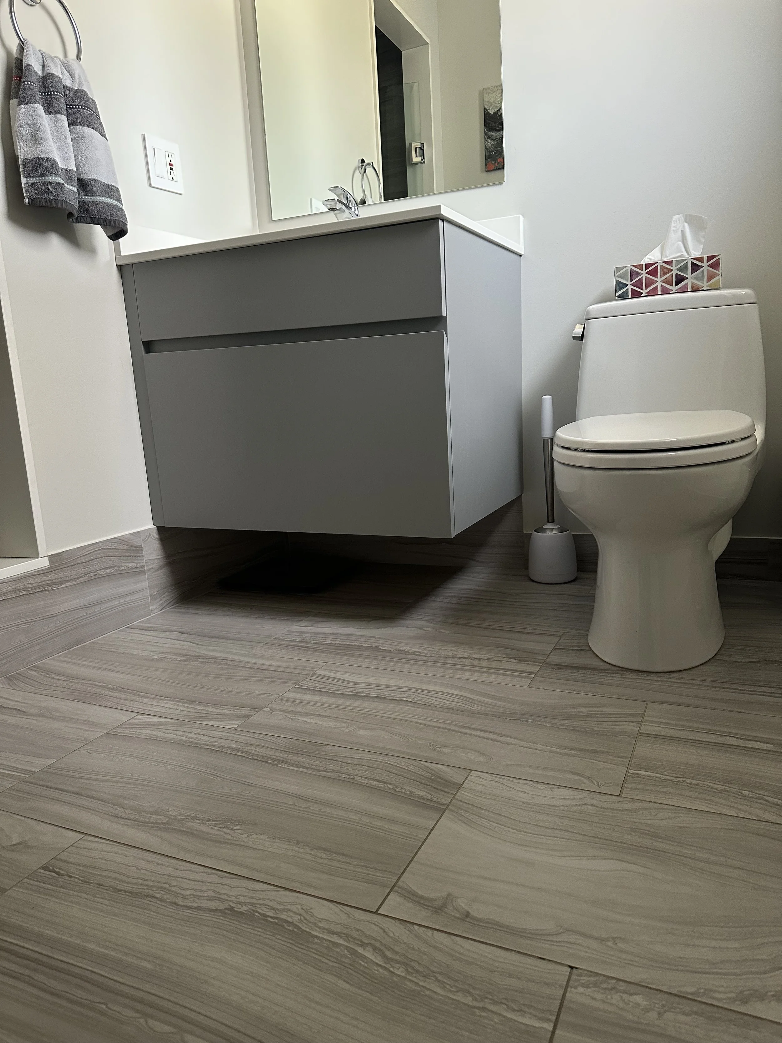 A bathroom with a white toilet, a grey vanity with a mirror, a grey and white towel, and a tissue box on the toilet tank.