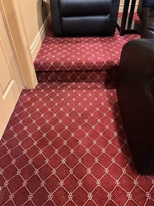 Red patterned carpet with a step-up platform in the corner of a room, showing part of a black leather armchair and furniture.