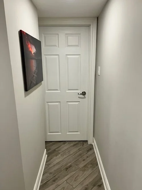 A narrow hallway with gray walls and wood flooring leading to a closed white six-panel door, with a black-and-red framed artwork on the left wall.