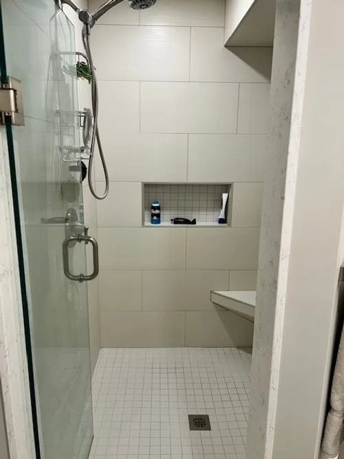 A modern shower and toilet area with a glass door, tiled walls, a built-in niche with toiletries, and a small drain on the white tiled floor.