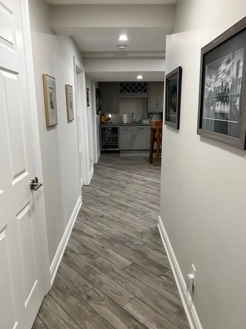 A hallway leading to a kitchen area with grey cabinets, a small wine rack, and a wooden barstool. The hallway has white walls with framed pictures and wood-look flooring.