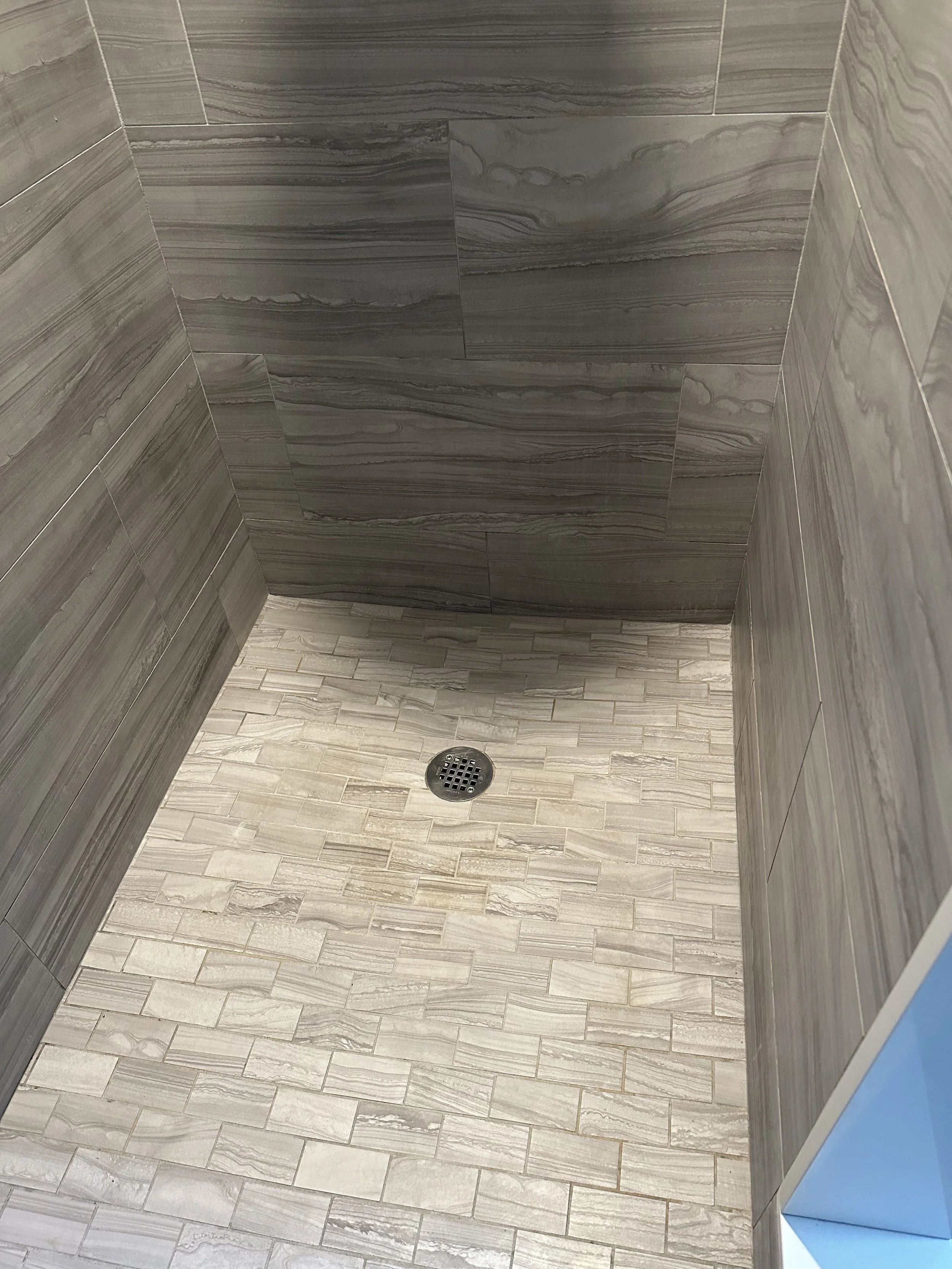 A walk-in shower with grey marble wall tiles and light grey floor tiles with a drain in the centre.