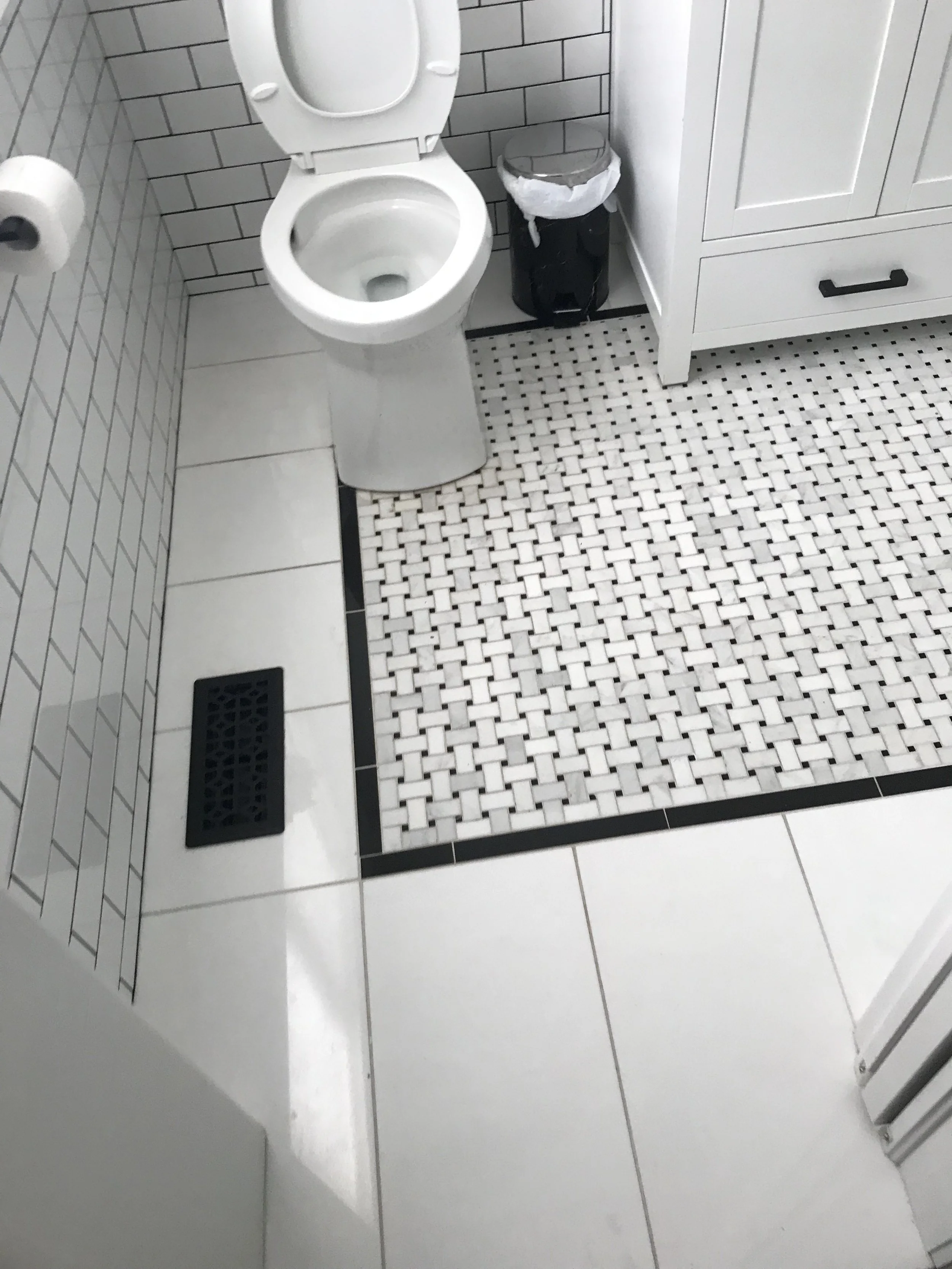 A bathroom with a white toilet, a black trash can with a plastic liner, a white cabinet, and a white tiled floor.