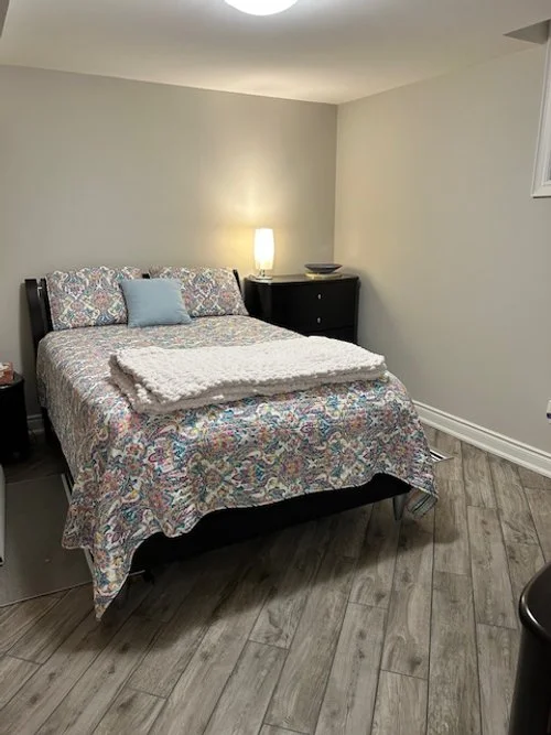 A bedroom with a bed, a black nightstand, a lamp, and a white blanket folded at the foot of the bed.
