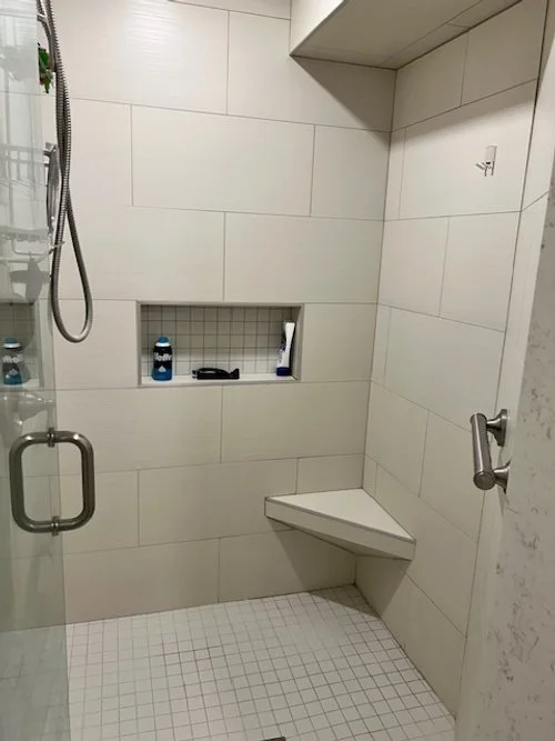 Shower stall with built-in niche holding shampoo bottles, razor, and body wash, tiled walls and floor, fold-down seat, and glass door.
