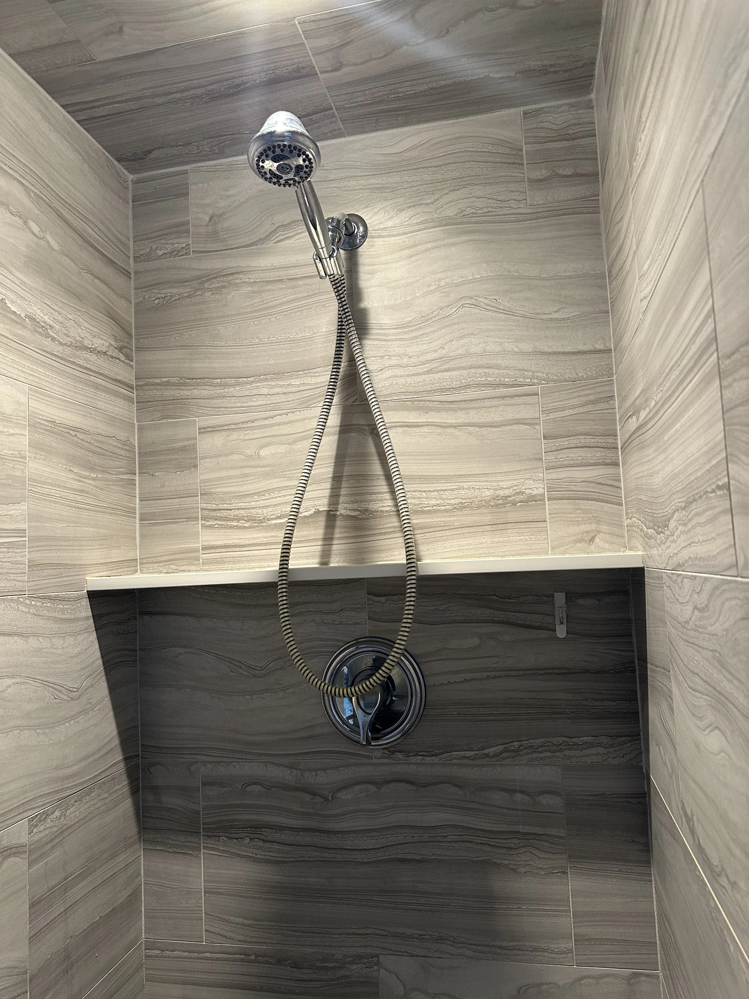 Shower with beige marbled tiles, a handheld showerhead, and a round water drain at the bottom.