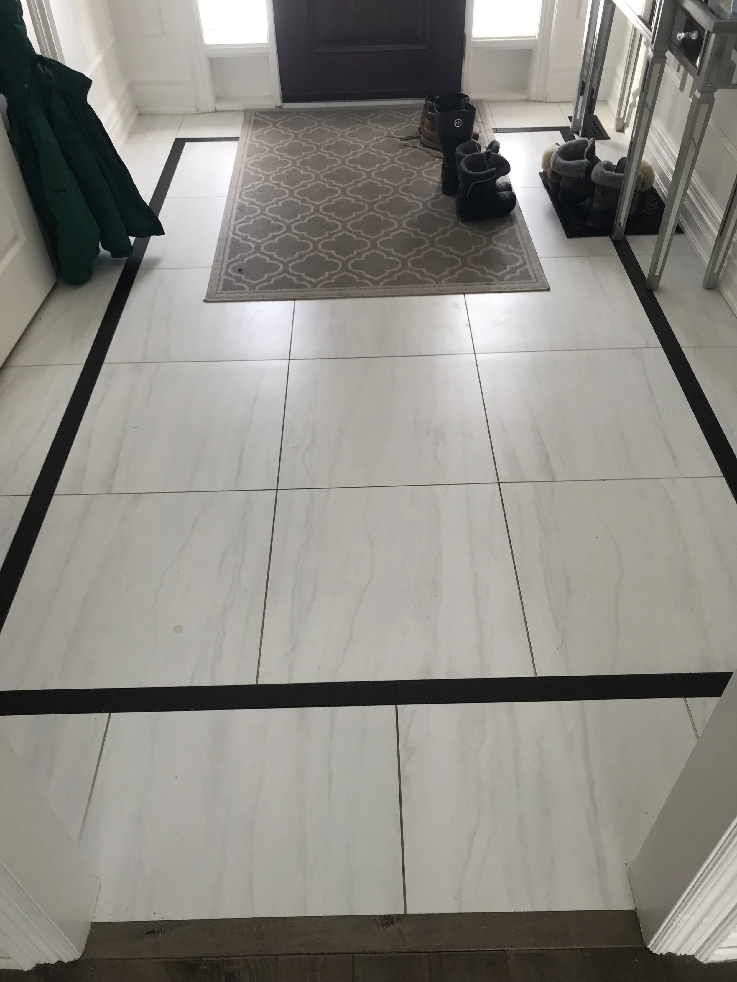 Entryway with large white tiled floor, black border accents, a small beige patterned rug near the black front door, and various shoes and boots on a shoe rack and the floor.