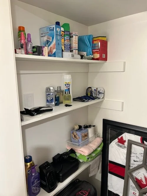 A small closet with three white shelves holding various personal care and household items, including lotions, sprays, a box, a framed photo, and a hockey jersey.