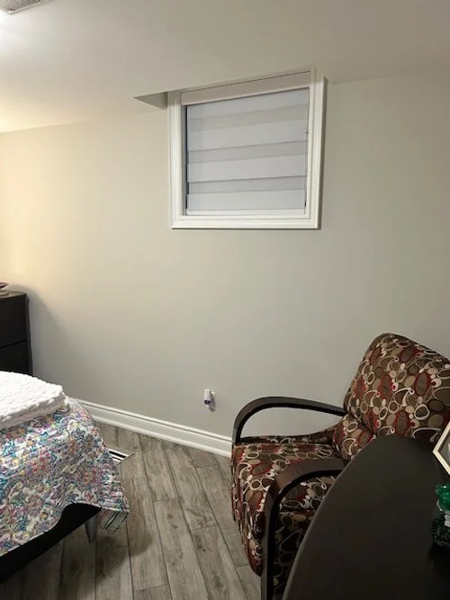 A small bedroom with a window, a patterned armchair, and part of a bed with a colorful quilt.