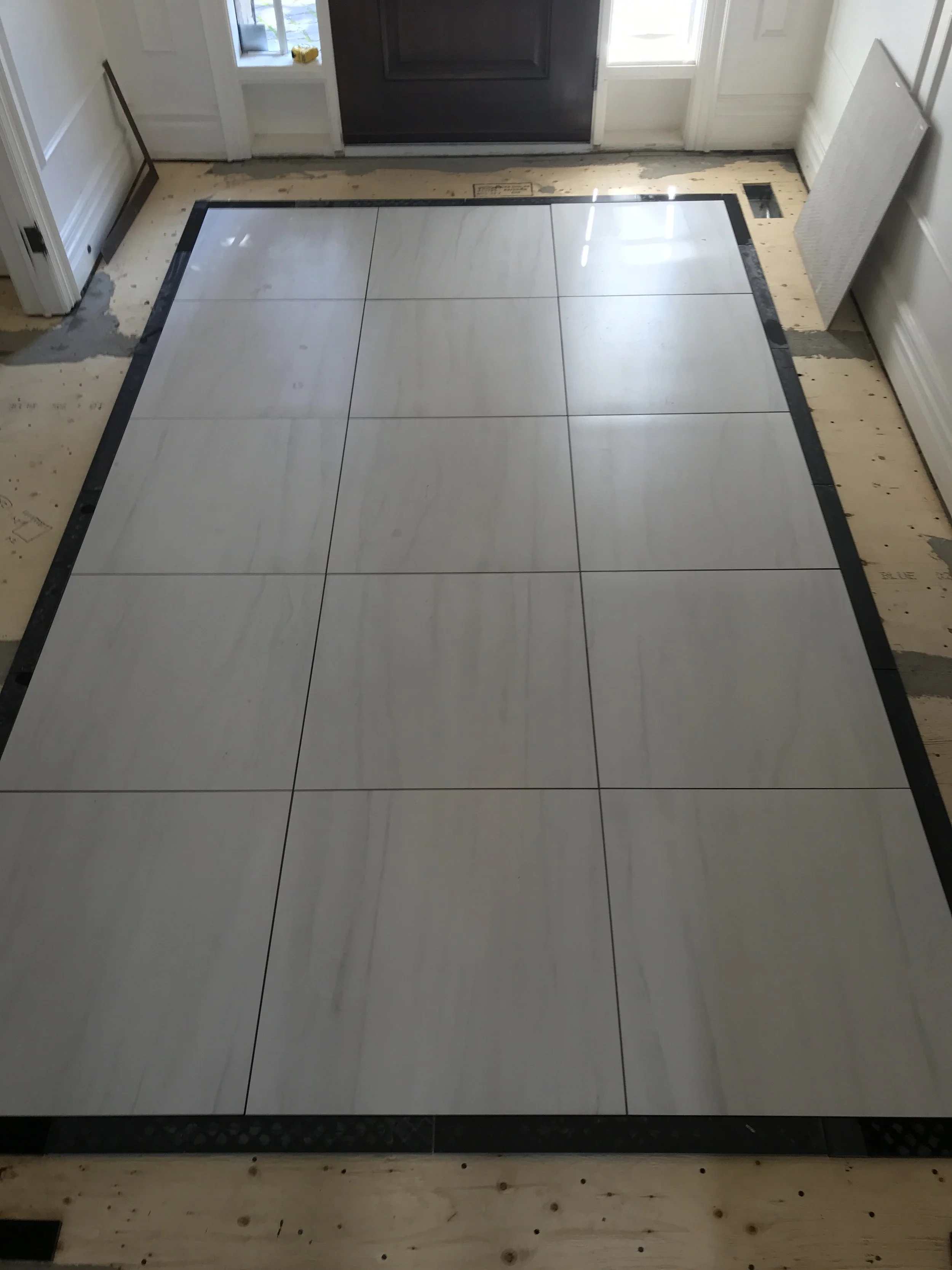 Newly installed large white tiles with subtle gray veining in a grid pattern on the floor of an entryway, with black border tiles around the perimeter.