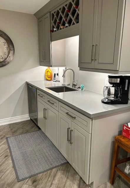 Kitchen with grey cabinets, white countertop, stainless steel sink, black coffee maker, decorative yellow bottle, wall clock, wine rack above cabinets, grey rug, and wooden side table.