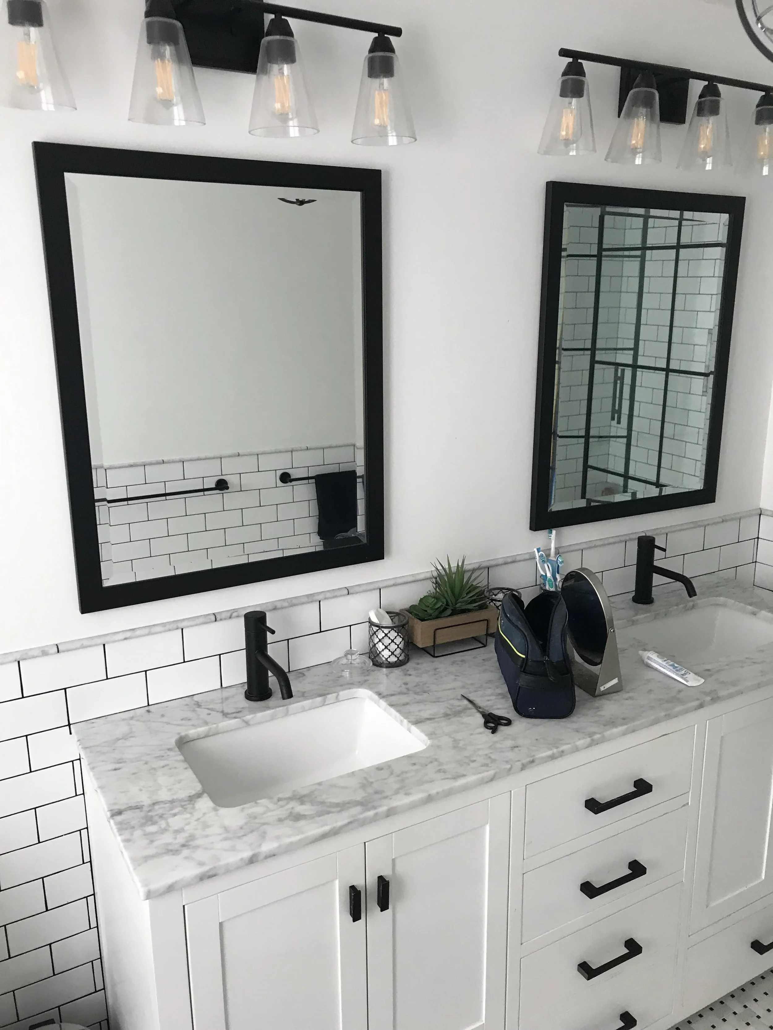 Modern bathroom vanity with white cabinet, marble countertop, and two black-framed mirrors. Items on the counter include a small plant, toothbrushes in a holder, scissors, a small speaker, and a remote control. Above are stylish light fixtures with clear glass shades.