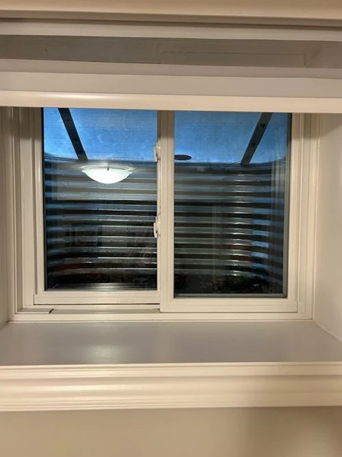 View through a window with white trim, showing a balcony with horizontal slats and a ceiling fan outside.