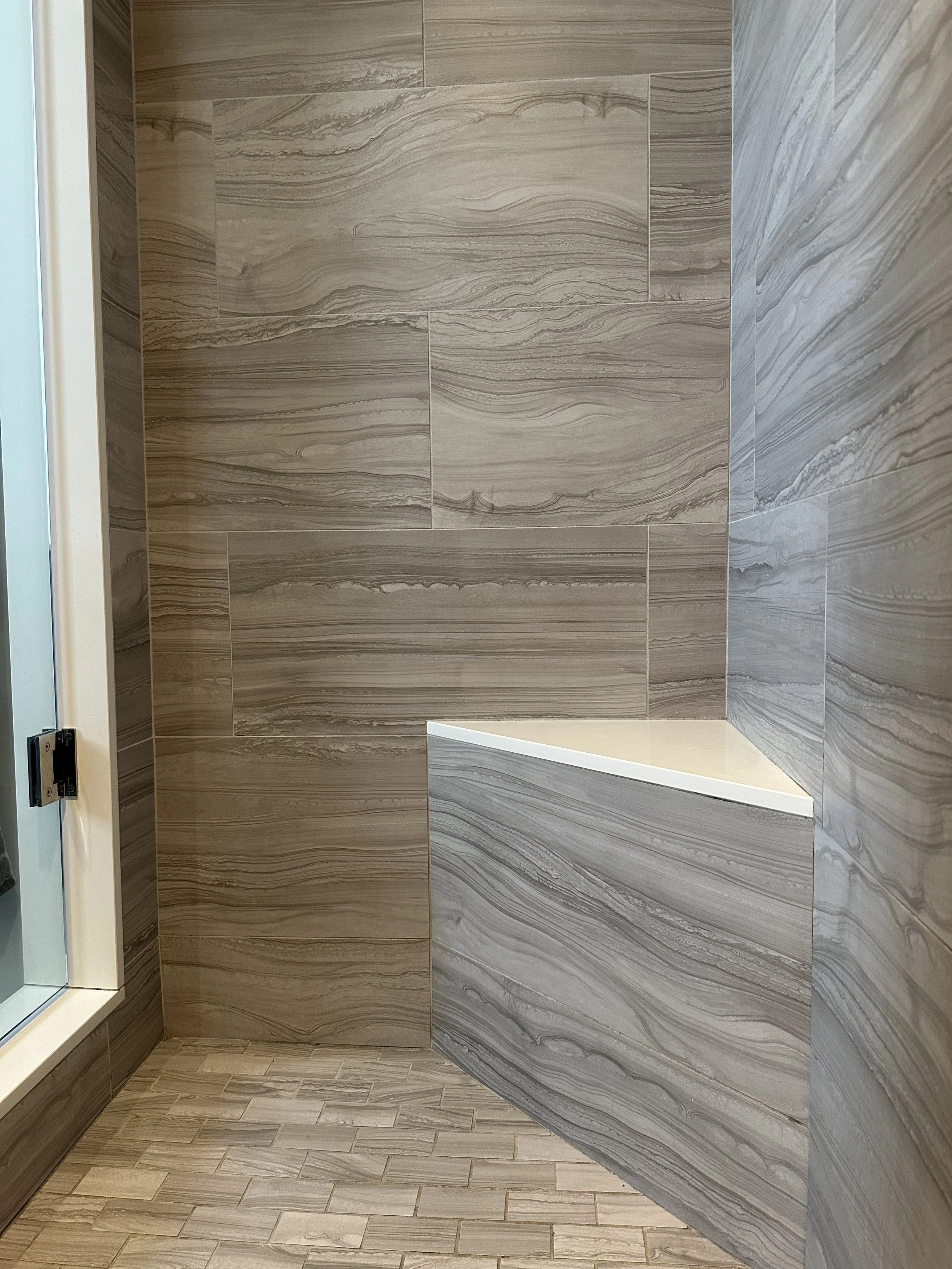 A bathroom shower with brown, marbled tiles on the walls and a built-in corner bench with a white top.