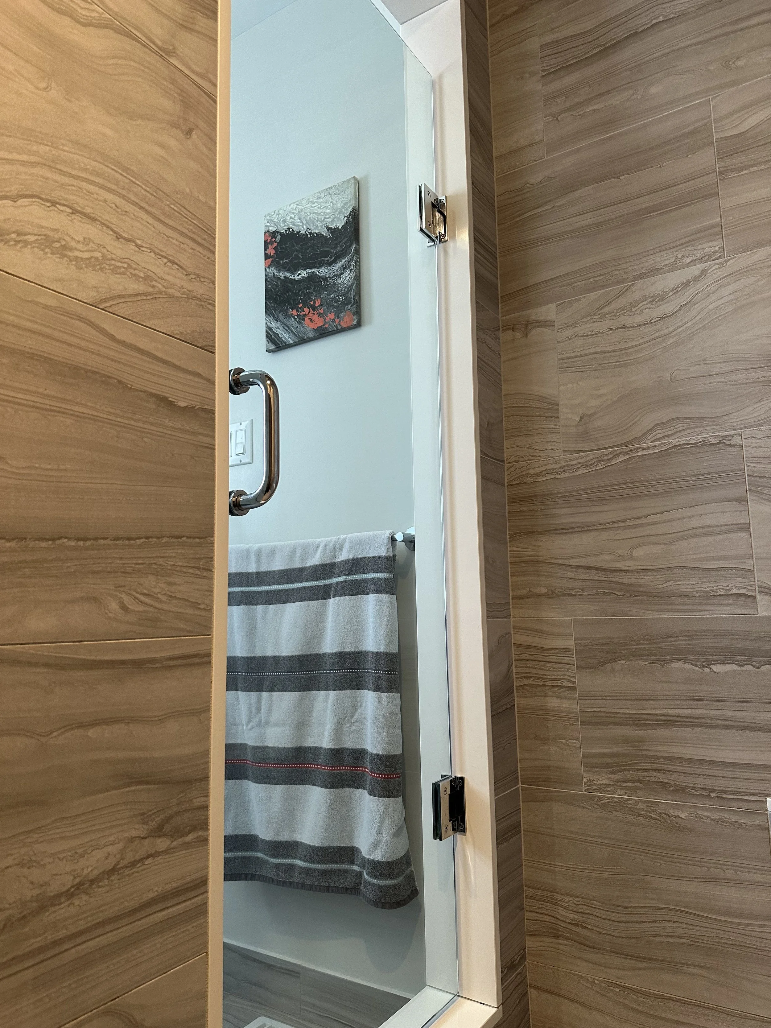 A bathroom shower with glass door, a towel hanging on a rack, and a painting on the wall.