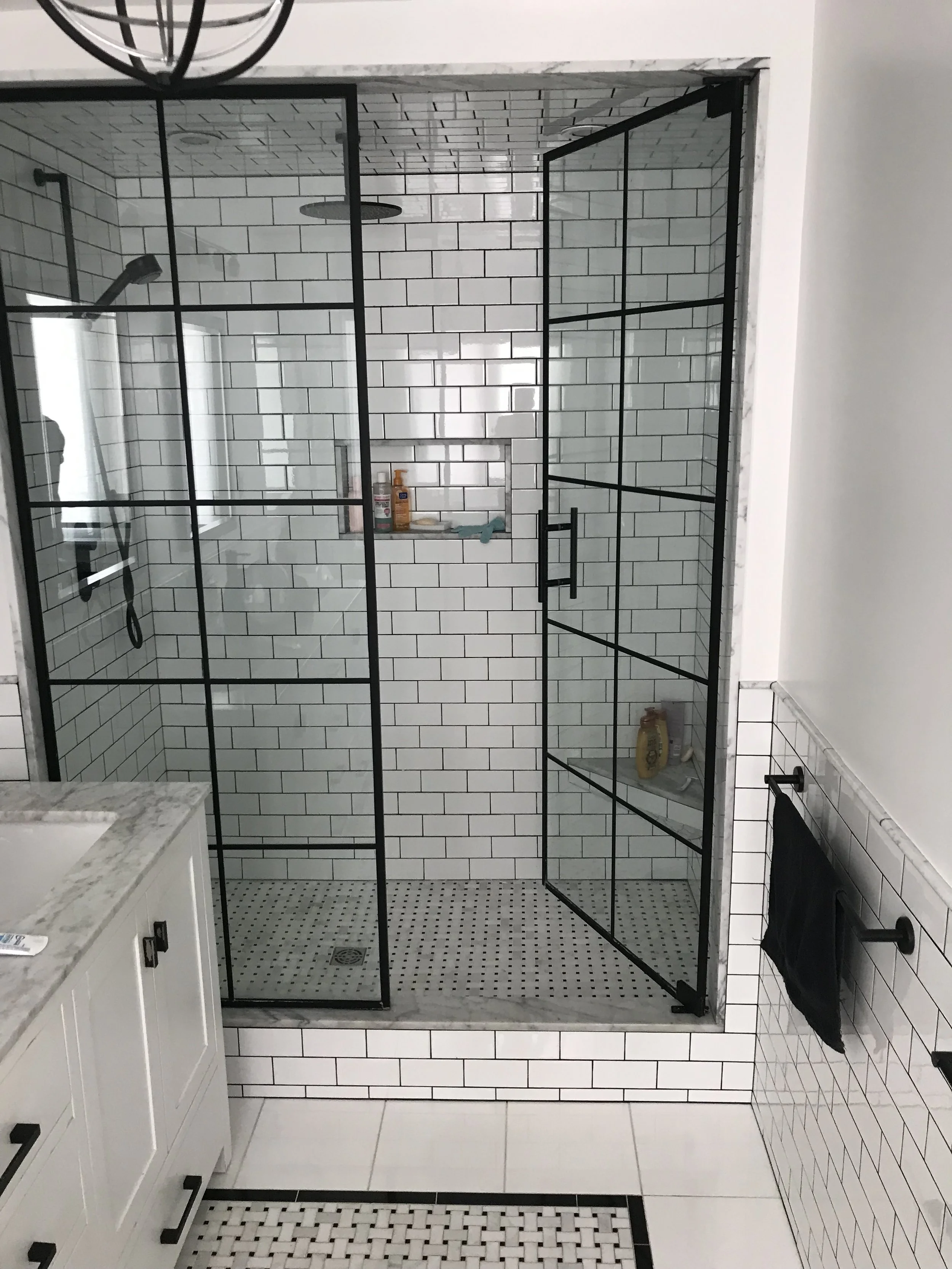 Modern walk-in shower with black metal framed glass door, white subway tiles, and built-in shelf holding toiletries, in a bathroom with white tiled walls and black accents.