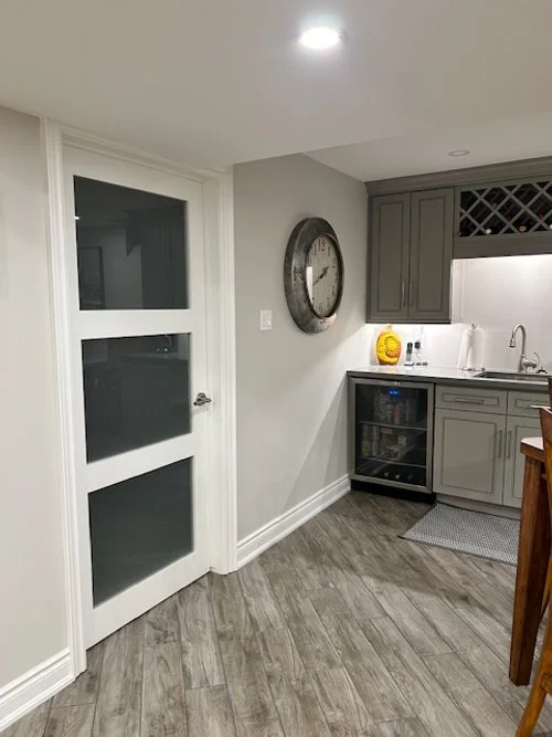 Image of a kitchen corner with grey cabinetry, a wine rack above the window, a clock on the wall, and a countertop with a small sink.