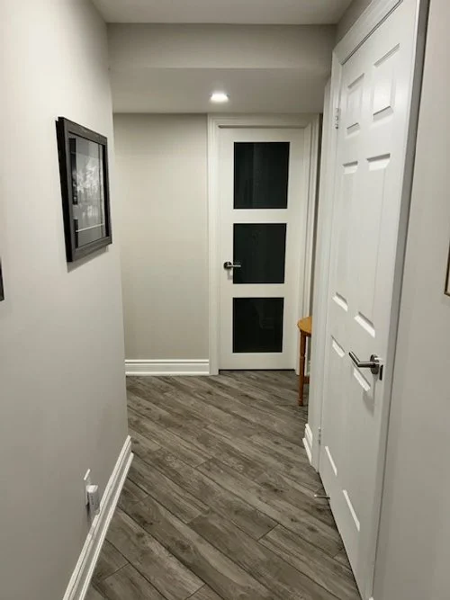 Empty hallway with white walls, wood flooring, and white doors, one with a silver handle, and a framed picture on the left wall.
