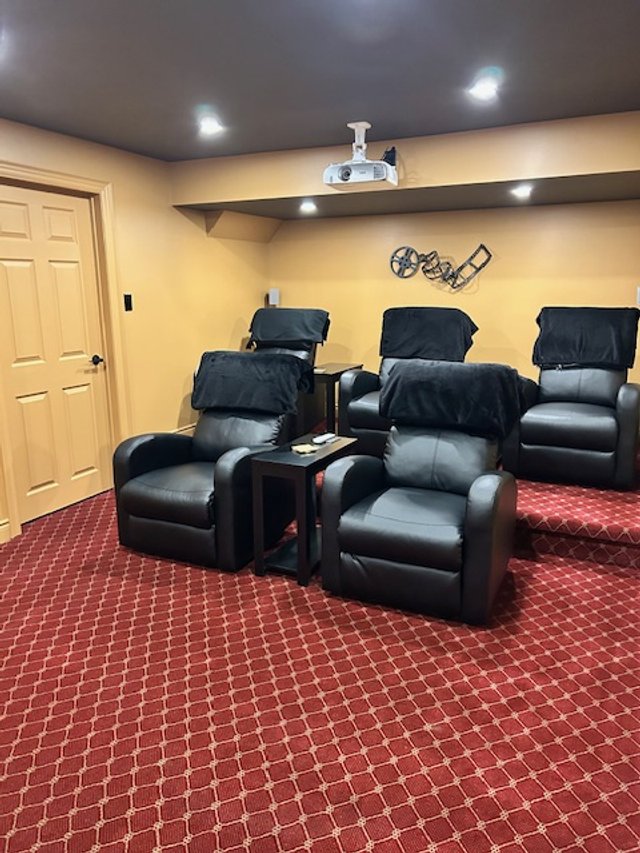Four black leather recliner chairs with black headrest covers, arranged in two rows in a home theater room with yellow walls and red patterned carpet, a small table ahead of each chair, and a ceiling-mounted projector.